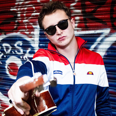 Liam C in a red, white and blue track top holding a guitar against a graffiti shutter